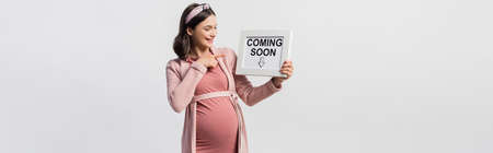 joyful pregnant woman pointing with finger at board with coming soon lettering isolated on white