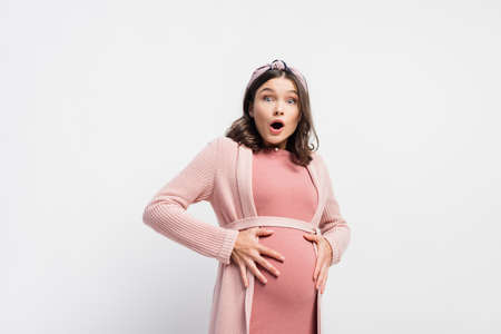 shocked and pregnant woman looking at camera and touching belly on white