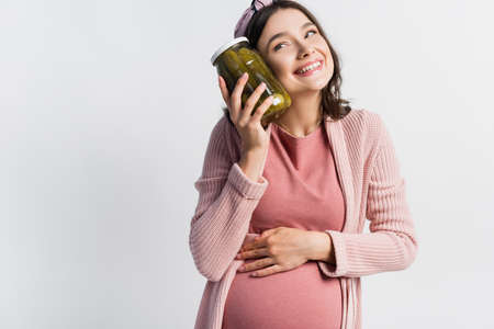 pleased and pregnant woman holding jar with pickled cucumbers isolated on whiteの写真素材