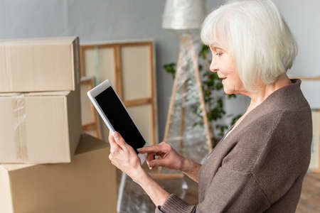 side view of senior woman holding digital tablet with blank screen with cardboard boxes on...