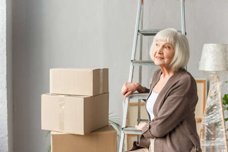 smiling senior woman sitting on ladder and looking away with cardboard boxes on background, moving conceptの写真素材