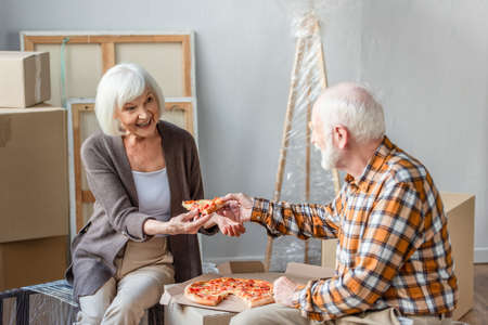 laughing senior man giving wife piece of pizza in new house and cardboard boxes on backgroundの写真素材