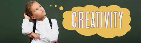 panoramic shot of thoughtful schoolgirl touching head and looking up near chalkboard with creativity...
