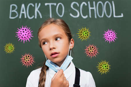 selective focus of exhausted schoolgirl taking off protective mask near chalkboard with back to school lettering and virus illustrationの写真素材