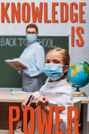 selective focus of schoolgirl in medical mask looking at camera while teacher standing near...