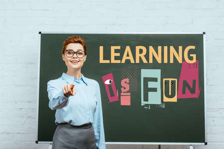 teacher looking at camera and pointing with finger near chalkboard with learning is fun lettering