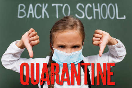 selective focus of displeased schoolgirl in medical mask showing thumbs down near chalkboard with inscription and quarantine lettering in classroomの写真素材