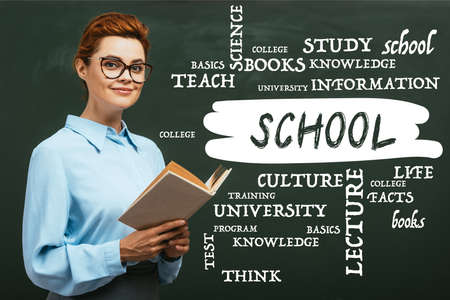 teacher in eyeglasses holding book near chalkboard with lettering in classroom
