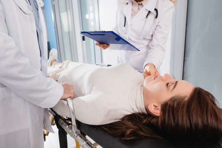 Selective focus of doctor with clipboard checking pulse of patient with closed eyes on stretcher...