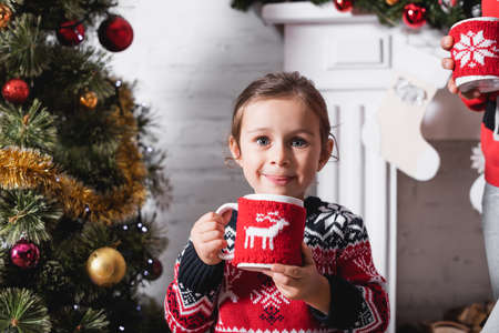 Front view of girl holding cup with knitted holder standing near festive pineの写真素材