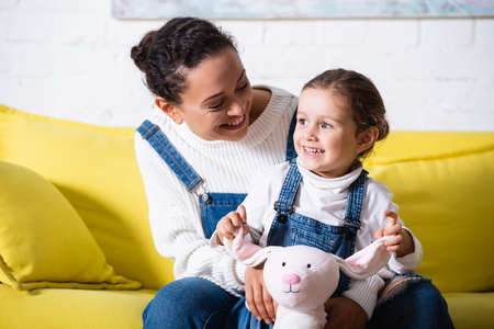 Mom sitting on couch and hugging daughter with soft toy at home