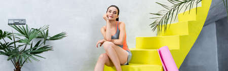 Panoramic orientation of sportswoman sitting on stairs near fitness mat at homeの写真素材
