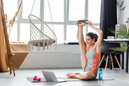 Sportswoman stretching leg while training near laptop on fitness mat at homeの写真素材