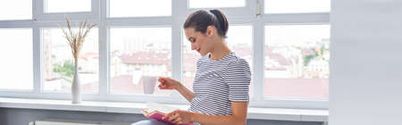Horizontal crop of woman with cup reading book near window at home