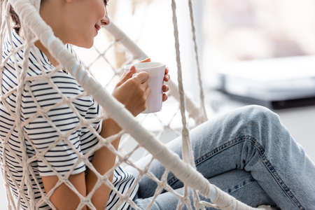 Cropped view of young woman holding cup in hanging armchair at homeの写真素材