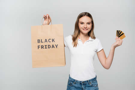 pleased woman holding credit cards and shopping bag with black friday lettering isolated on grayの写真素材