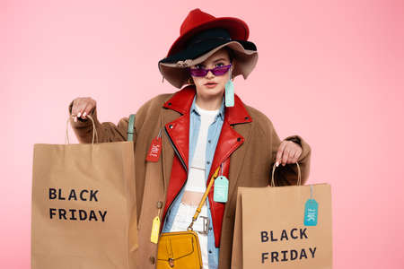 trendy woman in sunglasses and hats with sale tags holding shopping bags on pink, black friday...