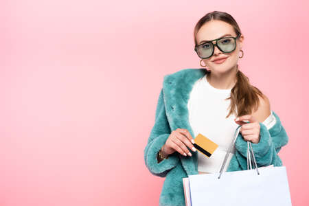 trendy woman in sunglasses holding credit card near shopping bag isolated on pink, black friday...