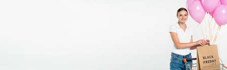panoramic crop of joyful woman holding shopping bag with black friday lettering near cart and pink balloons on whiteの写真素材