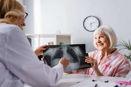 Elderly woman with pink ribbon and doctor holding chest x-ray at workplace