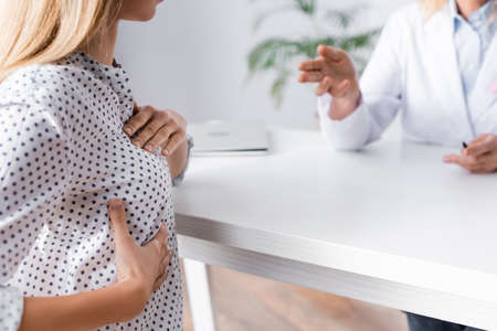 Selective focus of young adult woman with hands on breast sitting near doctorの写真素材
