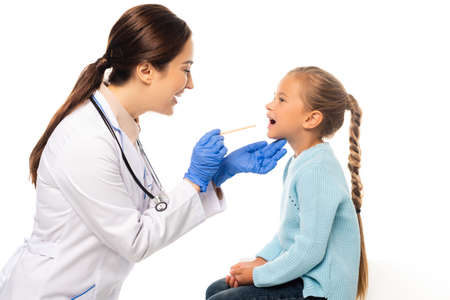 Smiling pediatrist holding stick near child with open mouth isolated on white