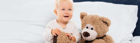 Panoramic shot of smiling toddler boy sitting near soft toy on bedの写真素材