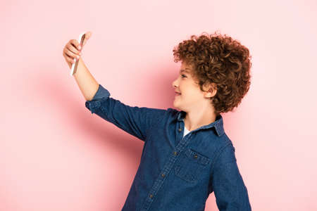 side view of curly kid in denim shirt taking selfie on smartphone on pinkの写真素材