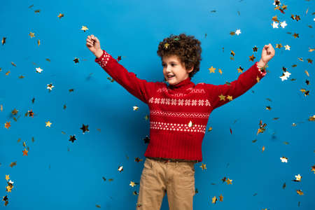 excited boy with hands above head in red sweater near golden confetti on blueの写真素材