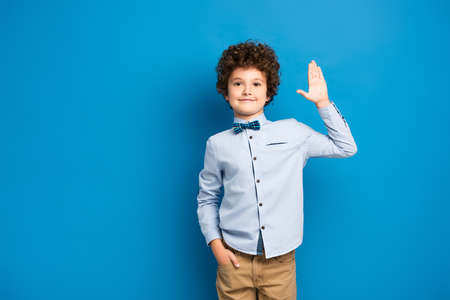 joyful kid in shirt and bow tie standing with hand in pocket and waving hand on blueの写真素材