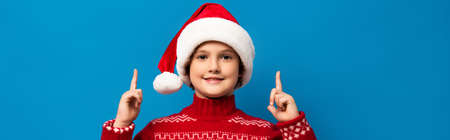 horizontal image of joyful kid in santa hat and red sweater pointing with fingers while looking at camera on blueの写真素材