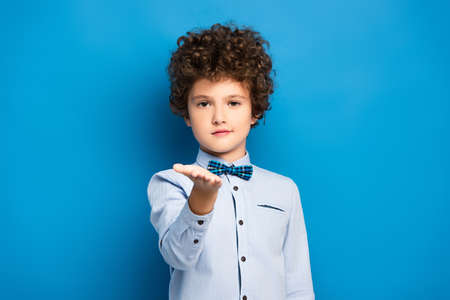curly kid in shirt and bow tie pointing with hand and looking at camera on blueの写真素材
