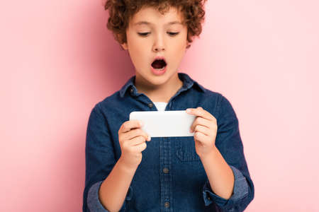 selective focus of surprised boy in denim shirt using smartphone on pinkの写真素材