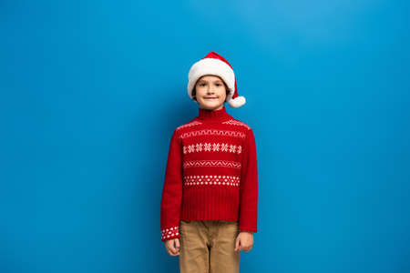 happy boy in santa hat and red knitted sweater looking at camera on blueの写真素材
