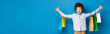 panoramic crop of joyful boy holding shopping bags and showing thumbs up on blueの写真素材