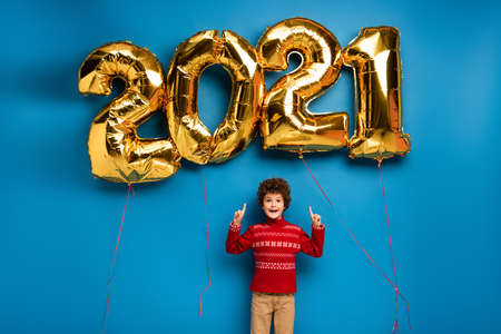 excited boy in red sweater pointing with fingers at golden balloons with 2021 numbers on blueの写真素材