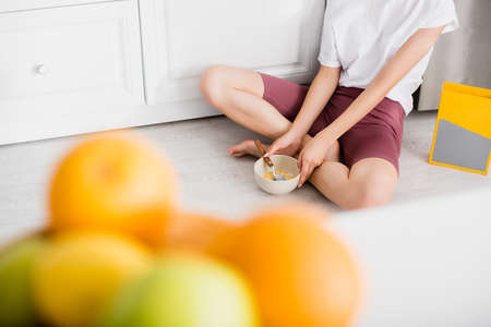 cropped view of young woman sitting on floor in kitchen with bowl of breakfast, selective focusの写真素材