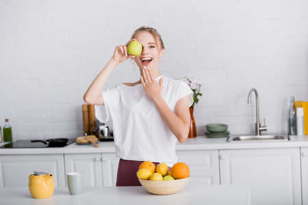 excited blonde woman touching chin while covering eye with fresh apple in kitchenの写真素材