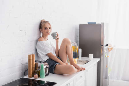 sensual young woman in shorts sitting on kitchen table and looking at cameraの写真素材