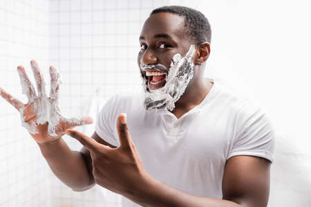 happy afro-american man with shaving foam on face pointing at handの写真素材