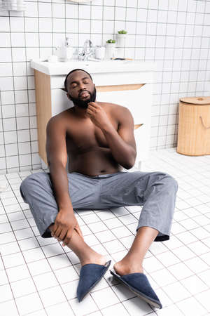 pensive afro-american man sitting on floor and touching beardの写真素材