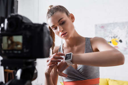 Selective focus of woman in sportswear pointing at fitness tracker near digital camera at homeの写真素材