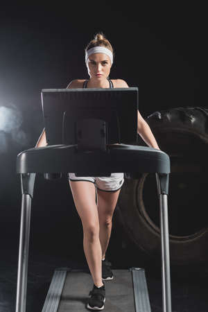Selective focus of young sportswoman training on treadmill near tire in gym with smoke