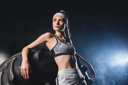 Young sportswoman looking away near tire in sports center with smokeの写真素材