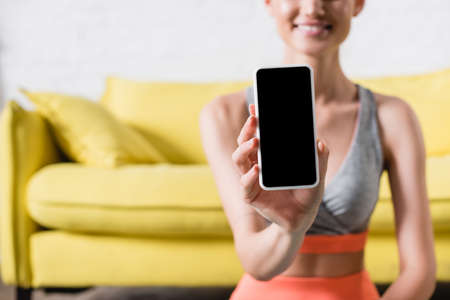 Cropped view of smiling sportswoman showing smartphone at camera in living roomの写真素材