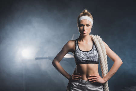 Sportswoman in headband with battle rope looking at camera in gym with smokeの写真素材