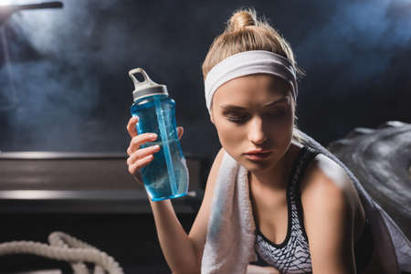 Selective focus of young sportswoman with towel holding sports bottle in gym with smokeの写真素材