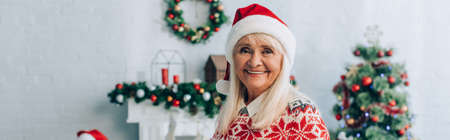 panoramic shot of happy senior woman in santa hat looking at camera near christmas tree on backgroundの写真素材