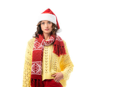 young brunette woman in santa hat and red scarf standing with hand in pocket isolated on white