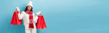 horizontal crop of joyful woman in winter outfit holding red shopping bags on blueの写真素材